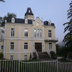 Large yellow historic building with steep dark roof, dormer windows, and a prominent central tower behind white picket fence.