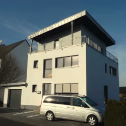 Modern three-story white house with balcony on top floor and a silver van parked in front.