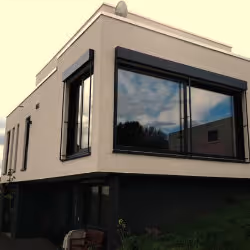 Modern two-story beige house with large corner windows reflecting the sky and nearby trees.